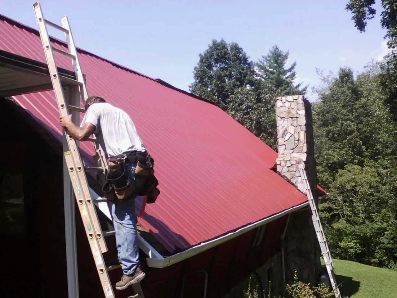 Roofer on a ladder beside a red metal roof and stone chimney — Soffit & Fascia Repair by Roofing Brothers in Shickley